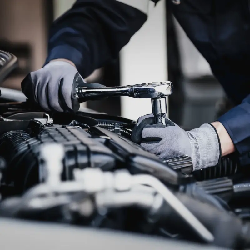 A gloved mechanic uses a chrome ratchet wrench on an engine under a car's open hood.