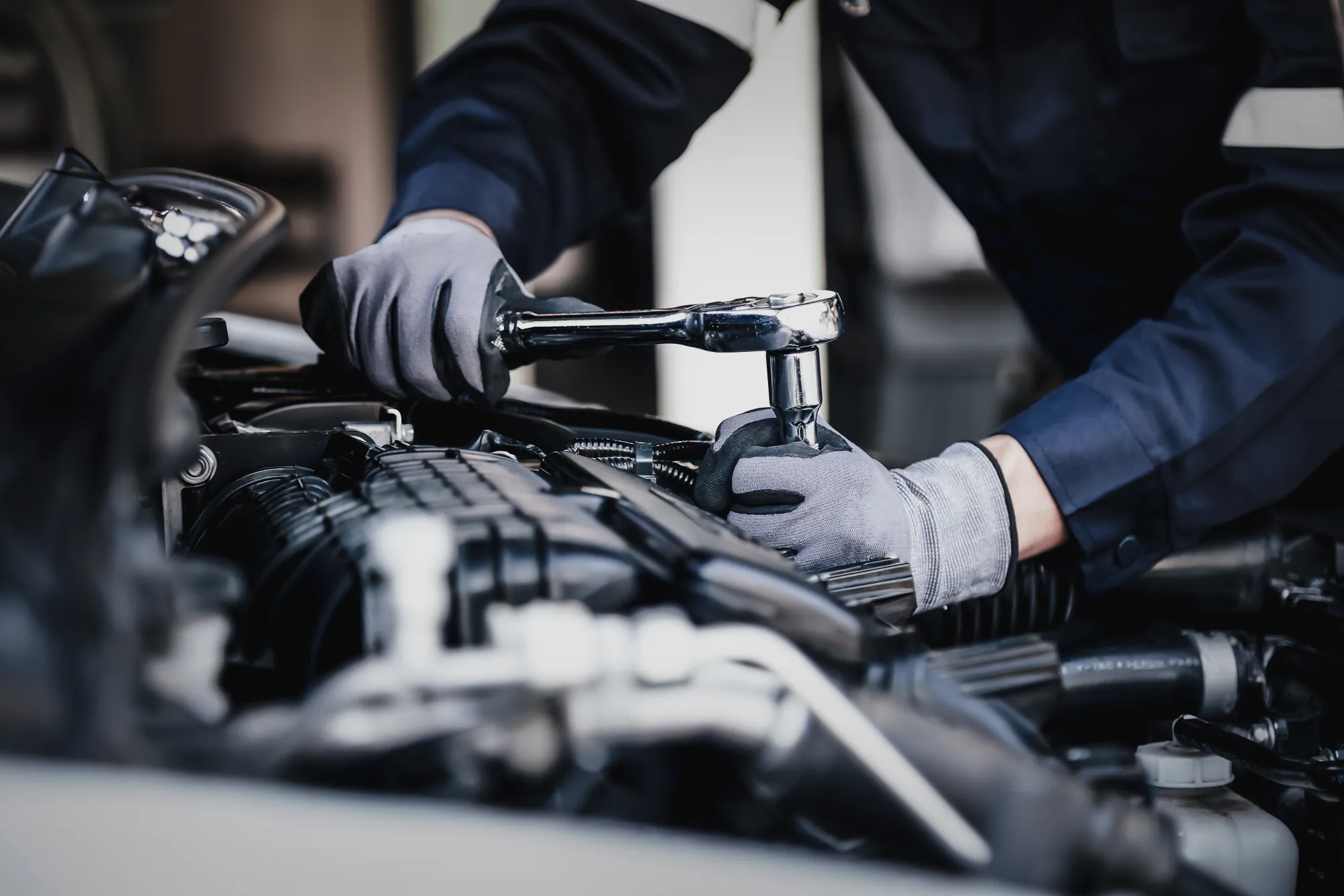 A gloved mechanic uses a chrome ratchet wrench on an engine under a car's open hood.